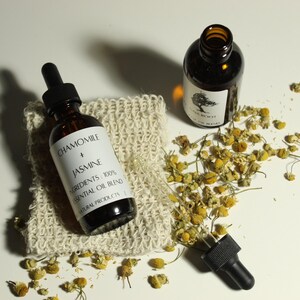 May include: Two amber glass bottles with black droppers, one labeled "Chamomile + Jasmine" essential oil blend. The bottles rest on a textured beige cloth and are surrounded by dried chamomile flowers. The image suggests a natural skincare or aromatherapy product.