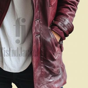 May include: A burgundy leather jacket with a button-up front and a pocket. The jacket has a classic design with a tailored fit. The sleeves have decorative detailing near the cuffs. The person is wearing a white shirt and blue jeans.