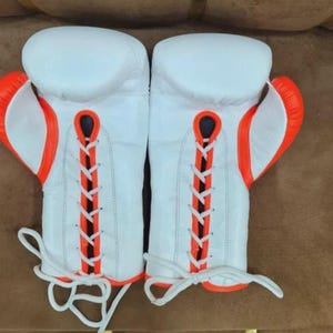 May include: Pair of white boxing gloves with orange accents. The gloves have a lace-up design with white laces and orange trim. The gloves are designed for boxing or other combat sports.