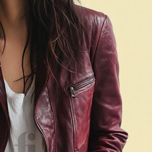 May include: A burgundy leather jacket with a zippered chest pocket and a silver zipper. The jacket is worn over a white top. The background is a solid light yellow.