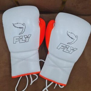 May include: A pair of white boxing gloves with orange accents. Each glove features a stylized bird graphic and the word "FLY" printed in black. The gloves have white laces.