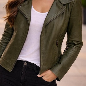 May include: Olive green leather jacket with a zipper closure, worn over a white top and black jeans. The jacket has a classic biker style with a notched collar and a small pocket.