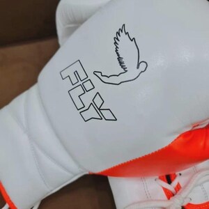 May include: White boxing gloves with orange trim and white laces. The gloves feature the word "FLY" and a graphic of a bird in black. The gloves are designed for boxing or other combat sports.