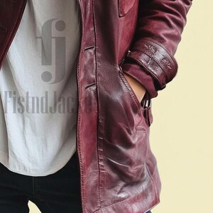 May include: A burgundy leather jacket with a buttoned front, side pockets, and a notched collar. The jacket has a tailored fit and features detailed stitching. The sleeves have decorative cuffs. The jacket is worn over a white shirt and blue jeans.