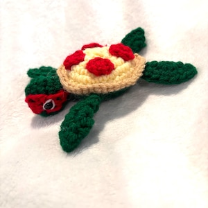 May include: A handmade crochet turtle with a pizza shell. The turtle has a green body, limbs, and head, with a red mask. The shell is beige with red pepperoni and a yellow cheese base. The turtle is lying on a white surface.