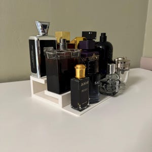 May include: A selection of perfume bottles in various shapes and colours, displayed on a white, tiered shelf. The bottles feature silver, gold, and black detailing, with some displaying text. The arrangement is on a white surface.