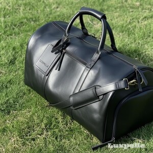 May include: A black leather duffel bag with a top handle and a shoulder strap. The bag has a zipper closure and a small tag with the word "Luxpelle" on it. The bag is sitting on green grass.