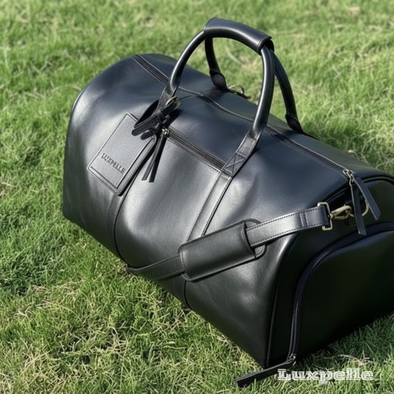 May include: A black leather duffel bag with a top handle and a shoulder strap. The bag has a zipper closure and a small tag with the word "Luxpelle" on it. The bag is sitting on green grass.