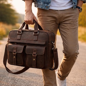 Könnte beinhalten: Eine braune Lederaktentasche mit zwei Vordertaschen, einem oberen Griff und einem Schultergurt. Die Tasche hat einen Vintage-Look mit sichtbaren Nähten und Metallbeschlägen. Die Aktentasche wird im Freien getragen.