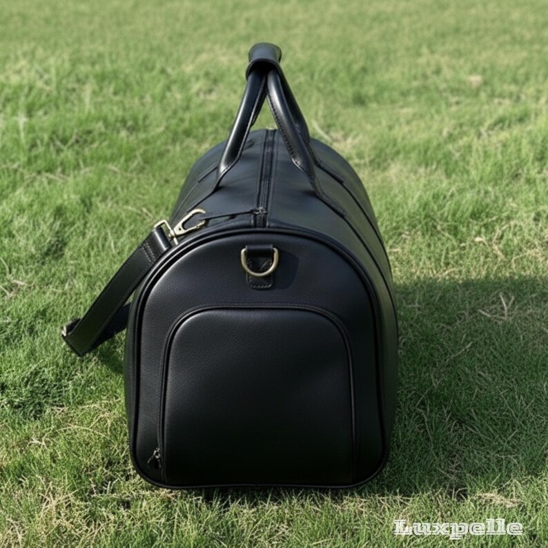 May include: A black leather duffel bag with a top handle and a detachable shoulder strap. The bag has a zippered main compartment and a front pocket. Gold-colored hardware accents the bag. The bag is sitting on green grass.