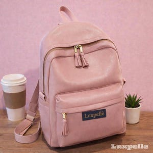 Op de afbeelding: Een roze rugzak met goudkleurige ritsen en kwastjes. De rugzak heeft een voorvak en een handvat aan de bovenkant. De merknaam "Luxpelle" staat op een blauw label. Een koffiebeker en een kleine plant staan ernaast.