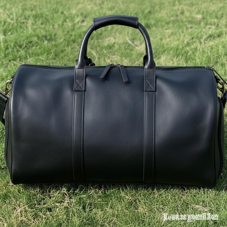 May include: A black leather duffel bag with a top handle and a detachable shoulder strap. The bag has a zipper closure and is sitting on green grass. The bag has a classic, minimalist design.