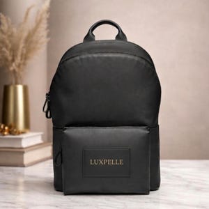 May include: Black backpack with a top handle and a front pocket. The front pocket features a rectangular label with the word "LUXPELLE" in gold lettering. The backpack is made of a smooth material and has a zip closure.
