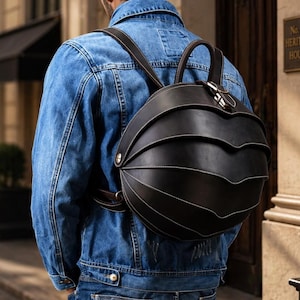 Puede incluir: Una mochila de cuero negro con un diseño único en capas y costuras blancas. La mochila tiene dos asas superiores y correas ajustables para los hombros. La persona de la imagen lleva una chaqueta vaquera azul.