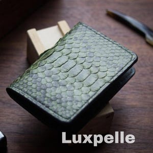 Może przedstawiać: Zielony portfel o fakturze skóry węża z białym napisem "Luxpelle". Portfel jest prostokątny i wydaje się być wykonany ze skóry. Tło stanowi ciemna drewniana powierzchnia.