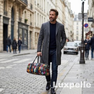 May include: A person walks along a cobbled street, holding a patterned duffel bag. They are wearing a grey overcoat, navy sweater, and blue jeans. The bag has a colourful cross design. The word "Luxpelle" is visible in the bottom right corner.
