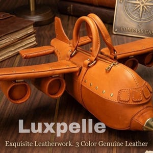 Op de afbeelding: Een handgemaakte, bruinleren handtas in de vorm van een vliegtuig met een ritssluiting, handvatten en klinknageldetails. De tas heeft vleugels en motordetails. De tekst "Luxpelle" is zichtbaar.