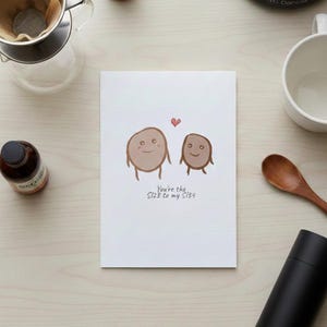 Op de afbeelding: Een witte wenskaart met een cartoon illustratie van twee lachende aardappelachtige figuren en een klein rood hart. De tekst luidt: "You're the S128 to my S134." De kaart ligt op een licht houten oppervlak.