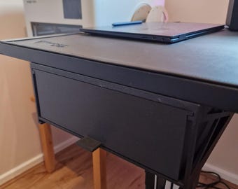 May include: A black adjustable desk with a drawer, a laptop, and a white printer. The desk has wooden legs and a black surface. A blue stylus rests on the laptop. The desk is in a room with wood flooring.