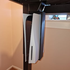 May include: A white and black gaming console mounted under a black desk. The console has a vertical design with a central black section and white side panels. The setup is against a light pink wall.