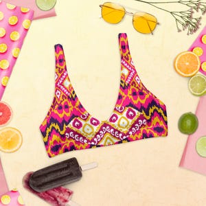 May include: A colorful bikini top with a vibrant geometric pattern in shades of pink, yellow, and purple. The top is a scoop neck style, perfect for swimwear. The image also includes sunglasses, fruit, and popsicles.