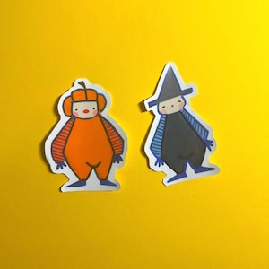 Könnte beinhalten: Zwei gestanzte Aufkleber mit stilisierten Figuren. Eine Figur trägt ein orangefarbenes Outfit im Kürbis-Design mit gestreiften Ärmeln. Die andere Figur trägt einen dunkelgrauen spitzen Hut und ein dunkelgraues Outfit mit blau gestreiften Ärmeln.