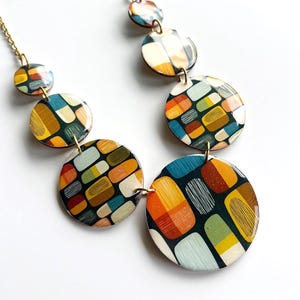 May include: A necklace featuring a gold-tone chain and multiple circular pendants of varying sizes. Each pendant displays a glossy, multi-colored pattern of rounded rectangular shapes in shades of orange, yellow, teal, green, and cream on a dark background.