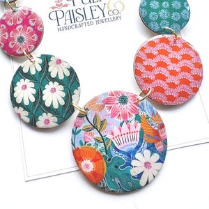 May include: A statement necklace with a gold chain and circular pendants adorned with floral patterns. The pendants showcase pink, teal, orange, and white designs. The necklace is handmade, with the brand name "Pulp & Paisley Co." displayed on a card.