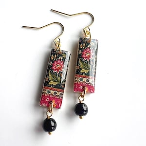 Könnte beinhalten: Ein Paar rechteckige Ohrhänger mit einem Blumenmuster in Rosa-, Grün- und Schwarztönen auf einer glänzenden Oberfläche. Jeder Ohrring hängt an einem goldfarbenen Haken und ist mit einer kleinen, runden, polierten schwarzen Perle versehen.