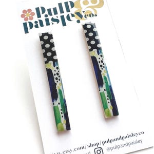 Peut inclure: Une paire de boucles d'oreilles rectangulaires en forme de barre avec un motif à pois noirs et blancs sur la moitié supérieure et un design abstrait de coups de pinceau multicolores en bleu, vert et jaune sur la moitié inférieure, sur un support.