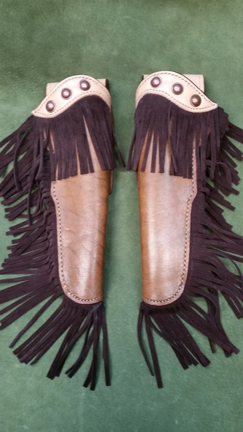 Op de afbeelding: Twee bruine leren holster met franjes. De holsters hebben een tan lederen bovenkant met decoratieve stiksels en metalen accenten.