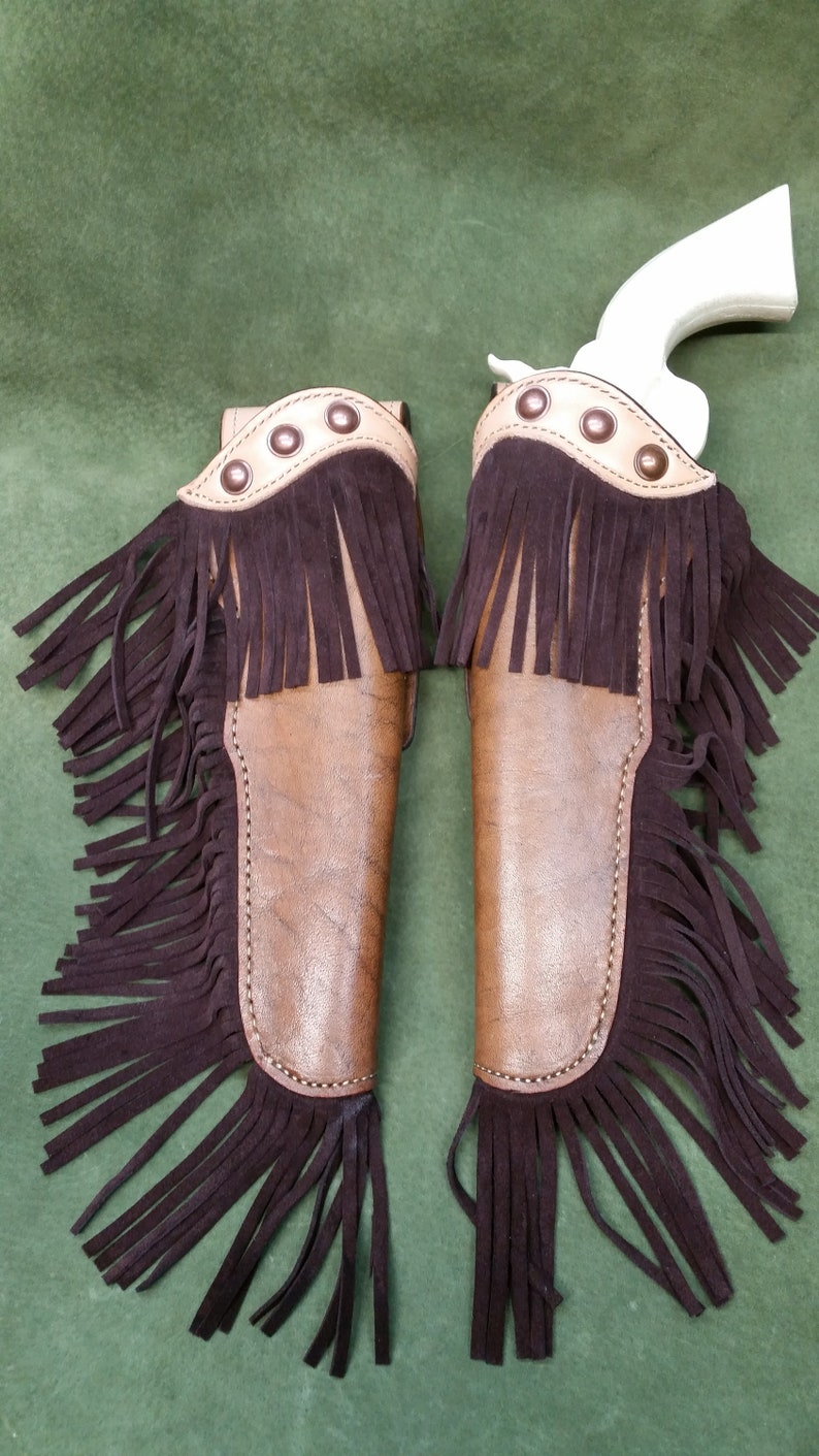 Op de afbeelding: Twee bruine leren pistoolholsters met franjes. De holsters hebben een tan lederen trim en metalen accenten. De franjes zijn bruin en hangen aan de onderkant van de holsters.