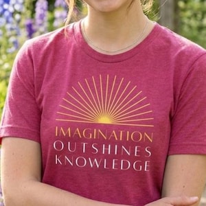 May include: A woman wearing a maroon t-shirt with the text "IMAGINATION OUTSHINES KNOWLEDGE" in gold lettering. The shirt features a sunburst design. The background is a garden with flowers.