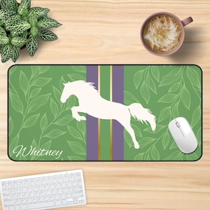 May include: A rectangular desk mat featuring a green background with white line-art leaves, a central purple vertical stripe with gold accents, and a white silhouette of a jumping horse. The name Whitney is printed in white script in the corner.