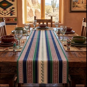 May include: A wooden dining table set for a meal, featuring a striped table runner in navy blue, green, and cream. The table is set with plates, bowls, glasses, and decorative figurines. The background includes a window with a desert landscape.