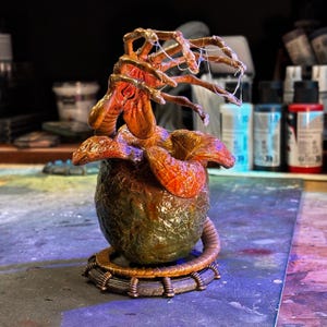 Op de afbeelding: Gedetailleerde sculptuur van een alien-ei met een opkomend wezen. Het ei is voornamelijk groen en bruin, met een textuur. Het wezen is oranje en bruin, met een complex, organisch ontwerp. De sculptuur staat op een cirkelvormige basis.