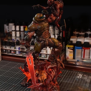 May include: A detailed figurine of a character in green and brown armour, holding a glowing red sword, battling a red creature. The base is a detailed, textured, and bloody scene. The figurine is set against a blurred background.