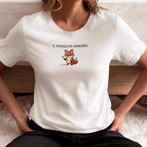 May include: White t-shirt with the text "i require snacks" above a cartoon fox illustration. The fox is holding a knife and is brown, white, and orange. The shirt is a crew neck and has short sleeves.