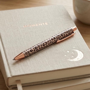 Könnte beinhalten: Ein beigefarbenes, stoffbezogenes Notizbuch mit dem Wort MOMENTE sowie einer Mondsichel und Sternen in Roségold-Prägung. Ein einziehbarer Kugelschreiber mit braun-schwarzem Leopardenmuster und roségoldenen Metallakzenten liegt oben auf.