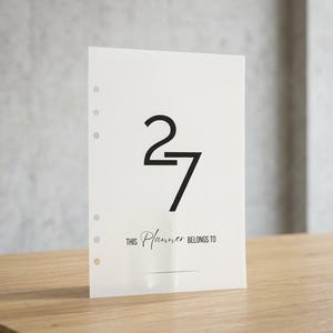 Könnte beinhalten: Eine weiße Planer-Einlage mit der großen schwarzen Zahl 27 in der oberen Mitte. Unter der Zahl steht der Text THIS Planner BELONGS TO, gefolgt von einer horizontalen Linie. Der linke Rand hat fünf kreisförmige Löcher für Ringbuchmechaniken.