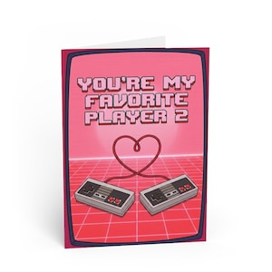 Puede incluir: Una tarjeta de felicitación con un tema de videojuego retro. La tarjeta presenta el texto "YOU'RE MY FAVORITE PLAYER 2" en fuente rosa pixelada. Dos mandos de juego grises están conectados por un cable rojo en forma de corazón sobre un fondo rosa.