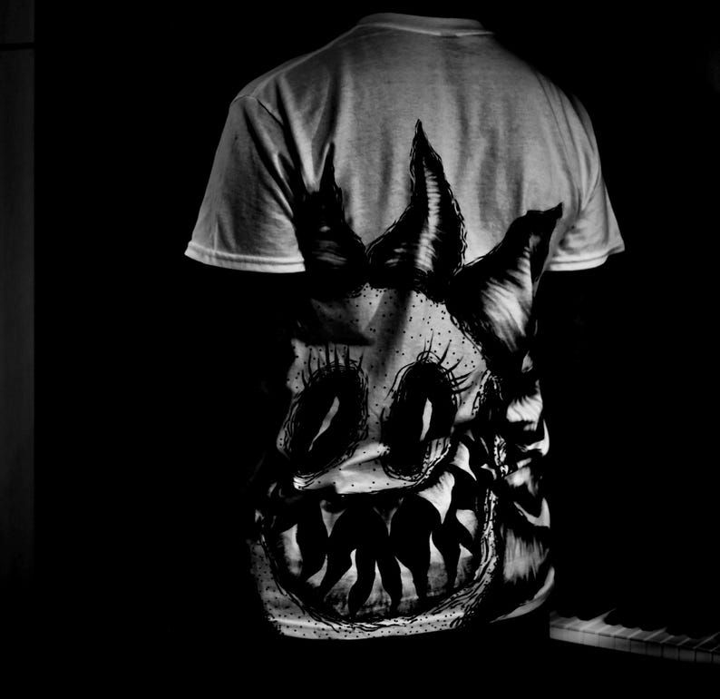 K&ouml;nnte beinhalten: Hellgraues T-Shirt mit einer schwarz-wei&szlig;en Grafik eines Monstergesichts. Das Design umfasst gro&szlig;e Augen, spitze Z&auml;hne und spitze H&ouml;rner. Die Grafik ist auf der R&uuml;ckseite des T-Shirts aufgedruckt.