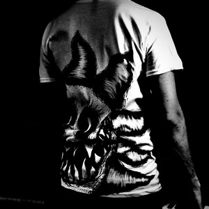 K&ouml;nnte beinhalten: Wei&szlig;es T-Shirt mit einer gro&szlig;en, detaillierten Schwarz-Wei&szlig;-Grafik eines Monstergesichts auf dem R&uuml;cken. Das Design zeigt scharfe Z&auml;hne und strukturiertes Fell oder Haut. Das T-Shirt wird von einer Person getragen, wobei R&uuml;cken und Arm sichtbar sind.