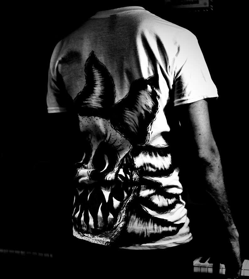 K&ouml;nnte beinhalten: Wei&szlig;es T-Shirt mit einer gro&szlig;en, detaillierten Schwarz-Wei&szlig;-Grafik eines Monstergesichts auf dem R&uuml;cken. Das Design zeigt scharfe Z&auml;hne und strukturiertes Fell oder Haut. Das T-Shirt wird von einer Person getragen, wobei R&uuml;cken und Arm sichtbar sind.