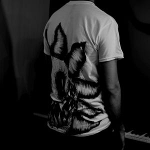 K&ouml;nnte beinhalten: Wei&szlig;es T-Shirt mit einem gro&szlig;en, detaillierten Schwarz-Wei&szlig;-Motiv auf dem R&uuml;cken. Das Design scheint ein stilisiertes, abstraktes Bild mit scharfen Kanten und einer dunklen, bedrohlichen &Auml;sthetik zu sein. Das T-Shirt wird von einer Person getragen, wobei die R&uuml;ckseite des Shirts zur Kamera zeigt.