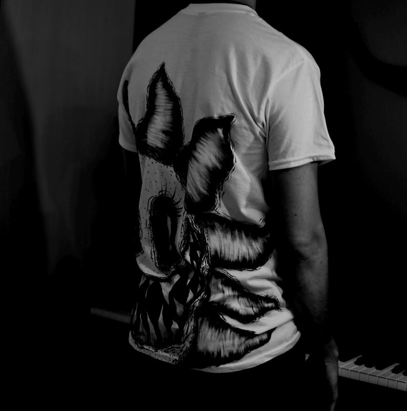 K&ouml;nnte beinhalten: Wei&szlig;es T-Shirt mit einem gro&szlig;en, detaillierten Schwarz-Wei&szlig;-Motiv auf dem R&uuml;cken. Das Design scheint ein stilisiertes, abstraktes Bild mit scharfen Kanten und einer dunklen, bedrohlichen &Auml;sthetik zu sein. Das T-Shirt wird von einer Person getragen, wobei die R&uuml;ckseite des Shirts zur Kamera zeigt.