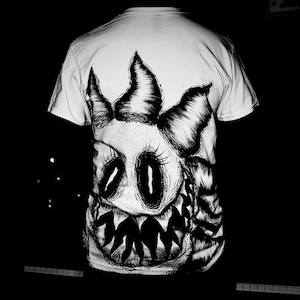 May include: White t-shirt featuring a detailed black and white illustration of a monster face. The design includes large eyes, sharp teeth, and a spiky crown. The artwork is centered on the shirt's front.