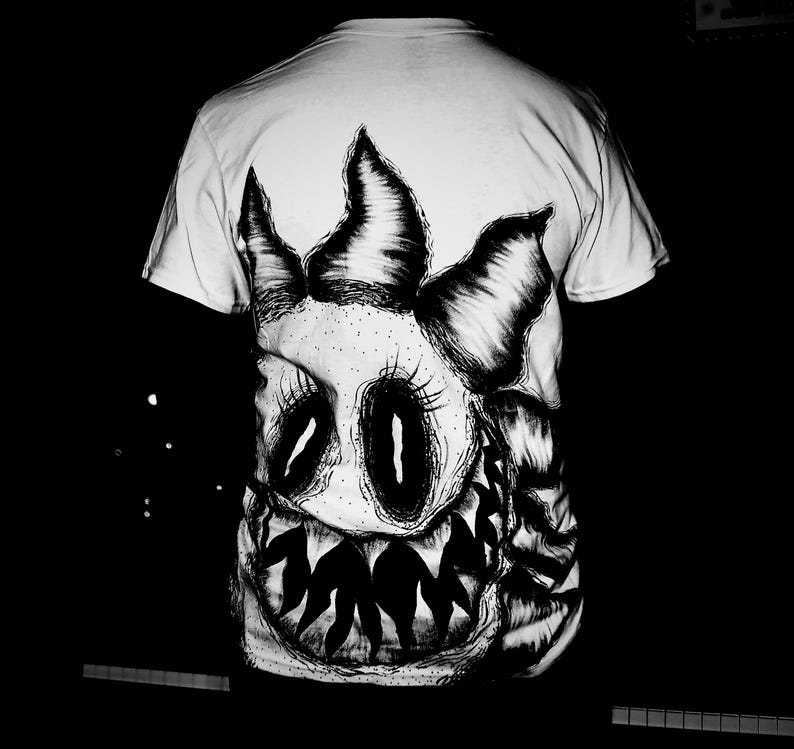 K&ouml;nnte beinhalten: Wei&szlig;es T-Shirt mit einer schwarz-wei&szlig;en Monstergesicht-Illustration. Das Design zeigt gro&szlig;e Augen, scharfe Z&auml;hne und eine stachelige Krone. Das Kunstwerk ist auf der Vorderseite des T-Shirts zentriert.