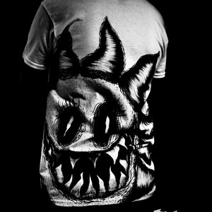 K&ouml;nnte beinhalten: Graues T-Shirt mit einer gro&szlig;en, detaillierten Schwarz-Wei&szlig;-Illustration eines Monstergesichts auf dem R&uuml;cken. Das Design umfasst spitze Z&auml;hne, gro&szlig;e Augen und spitze Ohren, was eine auff&auml;llige und kantige &Auml;sthetik erzeugt.