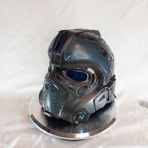 Puede incluir: Un casco futurista en tonos verde azulado y negro, con viseras y detalles en azul. El casco presenta paneles, ventilaciones y una base circular. El diseño sugiere un tema de ciencia ficción o cosplay.